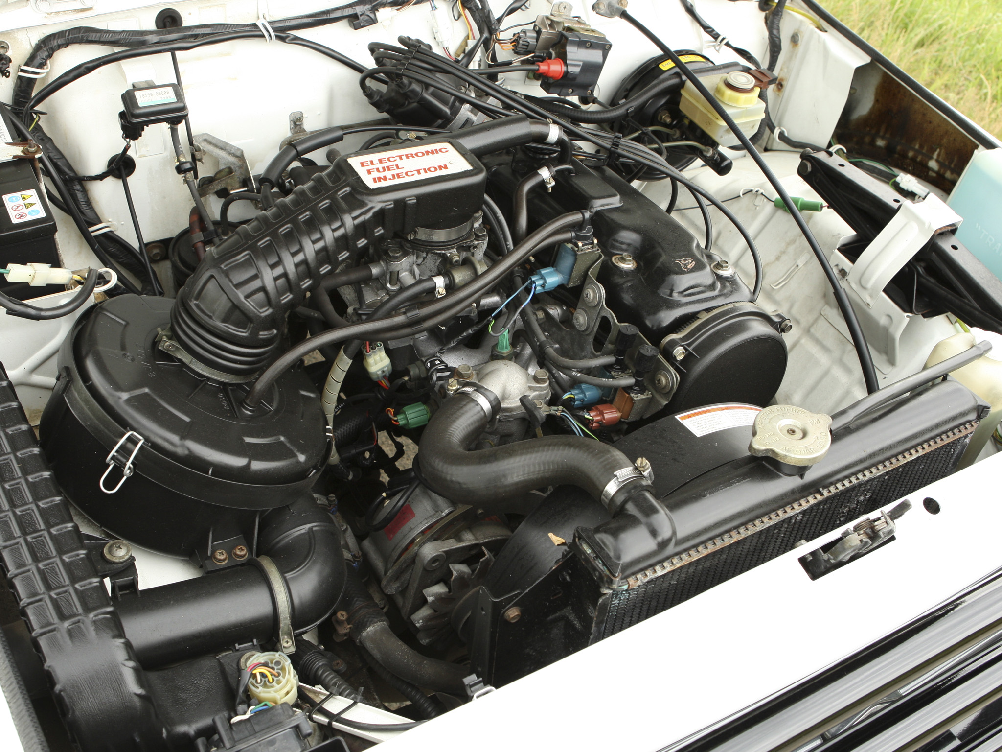 Под капотом Samurai Canvas Top (1990–95): G13BA с моновпрыском, крышка корпуса дросселя «ELECTRONIC FUEL INJECTION», трамблёр и катушка у щита; на пробке радиатора испанская надпись «APRETAR FUERTE / AFLOJAR LENTO» — вероятно, Santana 1.3i